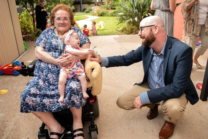 An elderly women in a wheelchair holding a baby girl while she looks towards her father smiling