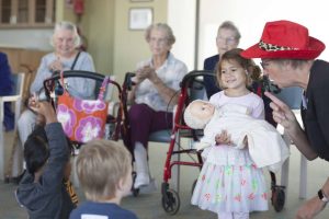 A young girl holding a baby doll smiling while elderly women look on happily