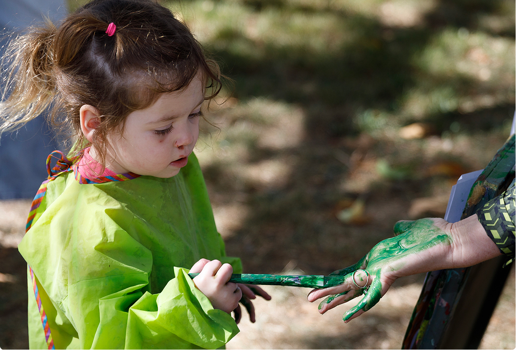 A little girl wearing a paint smock painting someones hand green