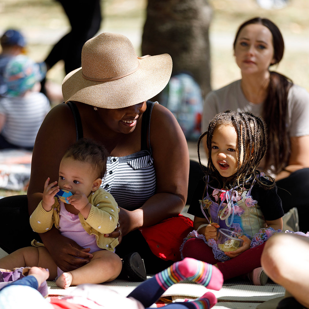 A mother and her children sitting on a rug outside engaged and smiling with others